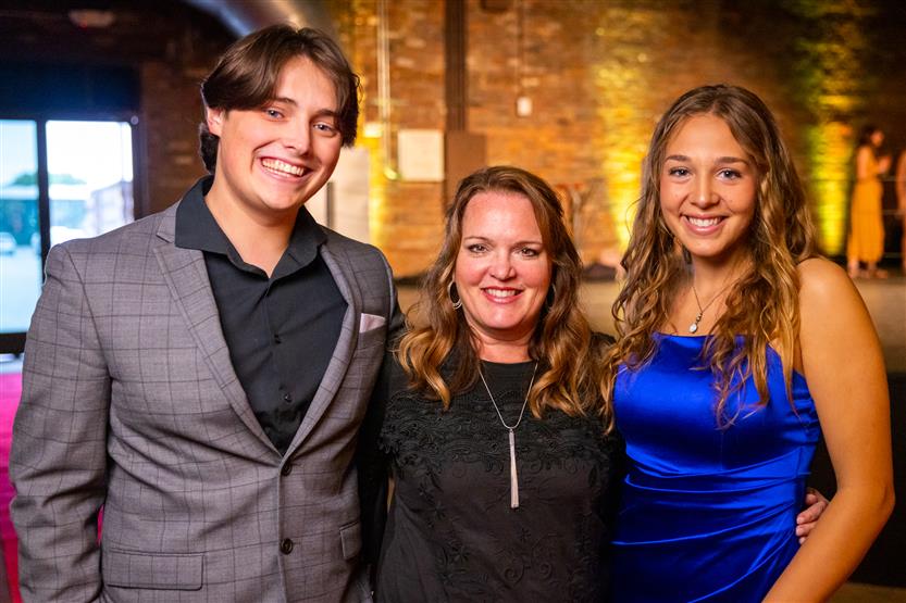 A young man wearing a gray checked blazer, a woman wearing a black dress and a woman wearing a blue dress smile